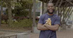 A man smiling outdoors with arms crossed, wearing a navy blue T-shirt with a gold logo.