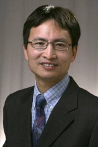 A professional portrait of a man in a suit and glasses, smiling for a business or news profile.