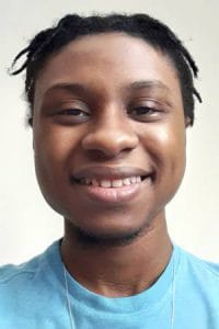 A smiling young man with dreadlocks wearing a blue shirt.