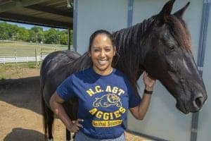 A woman in a blue N.C. A&T Aggies Animal Science t-shirt standing next to a black horse outdoors.