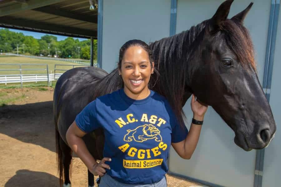 A woman in a blue N.C. A&T Aggies Animal Science t-shirt standing next to a black horse outdoors.