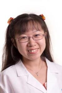 A woman with glasses and pigtails smiling, wearing a white lab coat, in a professional medical or sc.