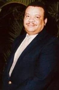 A person smiling in formal attire, standing outdoors with greenery in the background.