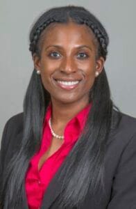 A woman in professional attire smiling, wearing a red blouse and black blazer, with a pearl necklace.