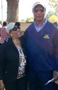 A woman and man smiling outdoors at a community event, wearing casual clothing and sunglasses.