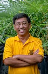 A man smiling in a yellow polo shirt standing outdoors among green plants.