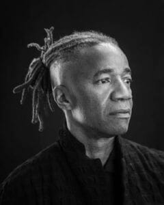 A woman with dreadlocks and a serious expression, wearing a dark top, in a black and white portrait.