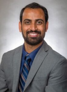 A professional headshot of a smiling man in a gray suit and blue tie, suitable for business or news.