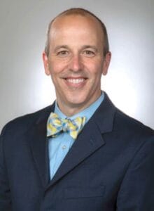 A man in a suit with a yellow bow tie smiling against a neutral background.