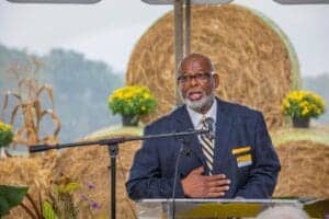 Agricultural expert speaking at outdoor event with hay bales and flowers in background.