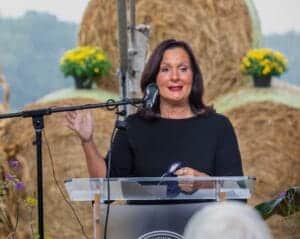 Woman speaking at outdoor event with hay bales and flowers in background.