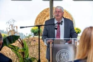 Agricultural expert speaking at outdoor event with hay bales and plants in the background.