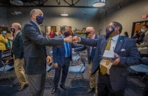 Business professionals greeting each other at a conference, wearing masks, in a modern meeting room.