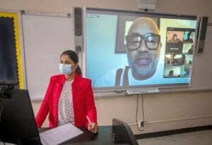 A woman in a red jacket and face mask participating in a virtual meeting in a classroom.