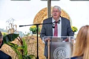 Agricultural expert speaking at outdoor event with hay bales and plants in the background.