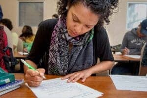Woman studying and writing notes in a classroom setting with other students.