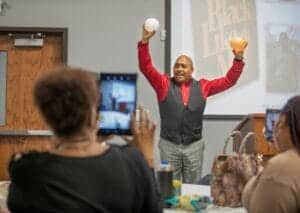 A woman giving a presentation with two balls, one white and one orange, in a classroom or conference.