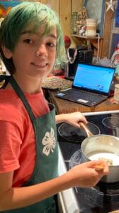 Young girl with green hair cooking on stove, wearing apron, in a cozy kitchen setting.