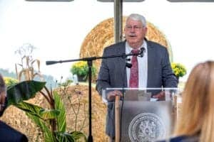 Agriculture official speaking at outdoor event with hay bales and plants in background.