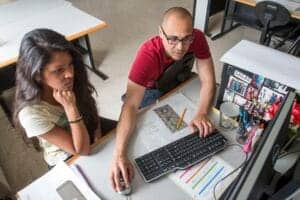A woman and a man working together at a desk with a computer and colorful magazines.