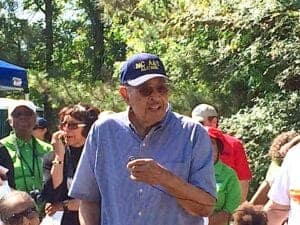An elderly man wearing a blue cap and shirt at an outdoor event with a group of people in a wooded a.