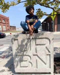 A young man sitting on a large concrete block with "YORBN" engraved, outdoors in an urban setting wi.