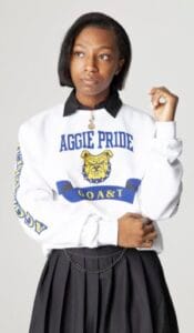 A woman wearing a white sweatshirt with "Aggie Pride" and a tiger logo, standing confidently.