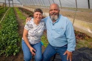 Healthy elderly couple smiling in a greenhouse with lush vegetable and flower beds.
