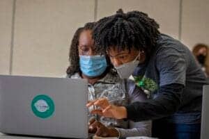 Accessible image showing two individuals working together on a laptop, both wearing masks, in a coll.