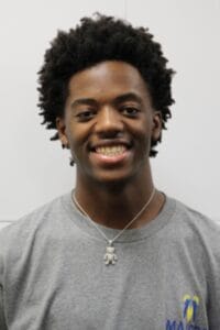 Young African American man smiling, wearing a gray T-shirt and necklace, in a professional setting.