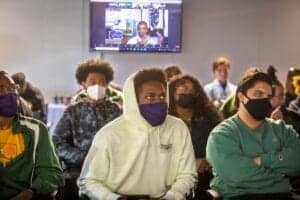Young diverse students wearing masks attending a classroom session with a virtual meeting displayed.