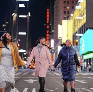 Brightly dressed women holding hands and laughing while walking in Times Square at night, illuminate.