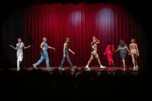 Diverse performers dancing on stage with red curtains at a cultural event.