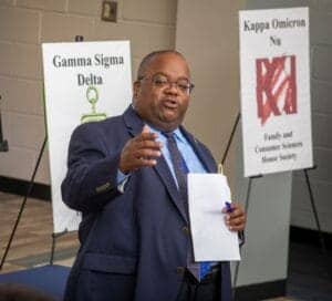Prominent man speaking at a professional event with fraternity banners in the background.