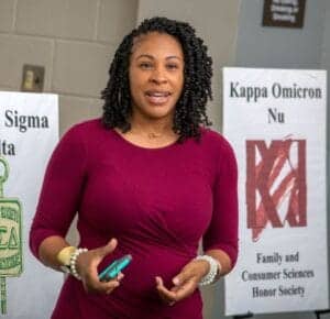 COVID-19 awareness speaker at CAES News event, woman in maroon dress with posters in background.