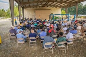 Community gathering at an outdoor event with a stage and audience under a pavilion.