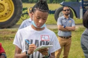 Young woman taking notes at outdoor CAES News event with farmers and machinery.