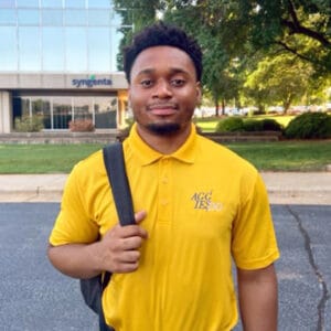Young man in yellow AGC TEXPO polo shirt holding backpack outdoors.