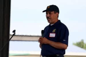 A man speaking into a microphone at an outdoor event, wearing a navy blue polo shirt and cap.