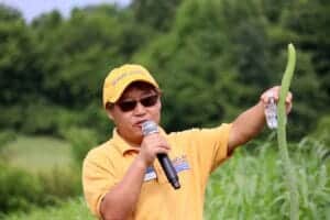 A woman in a yellow shirt and cap holding a microphone and a green plant in an outdoor agricultural.