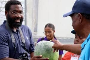 A man showing a large watermelon to a group of children outdoors.