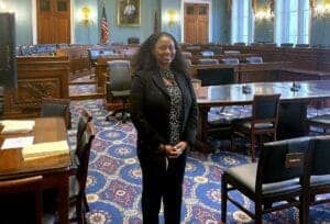 Accessible image description: A woman standing confidently in a formal government or legislative cha.