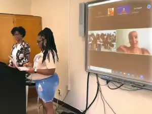 Students presenting in a classroom with a digital screen displaying a virtual meeting and participan.