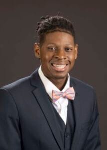 A young man in a navy suit and pink bow tie smiling confidently.