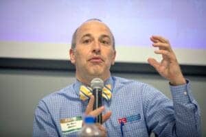 Expert speaker at conference wearing blue checkered shirt and yellow bow tie, holding a microphone d.