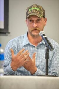 Man speaking at a microphone wearing a camouflage cap with "MOWING" text.