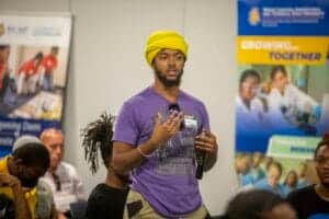 Young man speaking at a conference or seminar, holding a microphone and wearing a yellow headwrap.