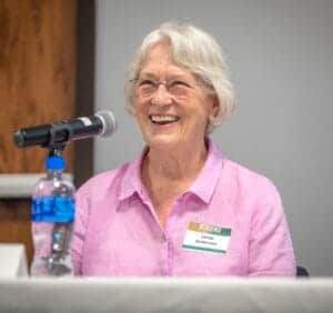 Bright smiling elderly woman with white hair and glasses, speaking at a conference or event, wearing.