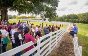 A colorful outdoor gathering of diverse people listening to a speaker on a farm or ranch setting.