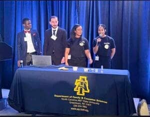 Students speaking at a panel on stage with a table displaying university logo and information.